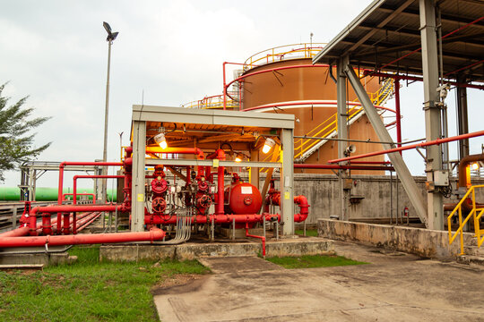 Industrial landscape showing a fire protection system with red pipes and pumps in front of massive oil storage tanks at a refinery