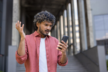 A worried young Indian man stands on the street outside an office building and stares in dismay at...