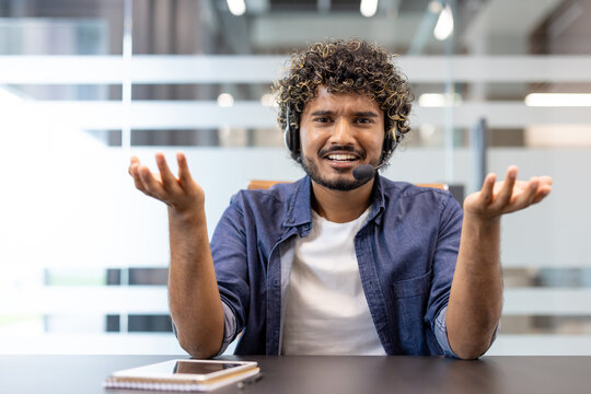 Dissatisfied young Indian man sitting in a headset at an office desk, talking and gesturing anxiously at the camera