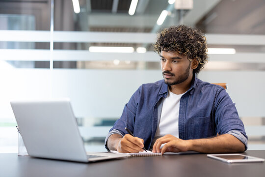 A young Indian man works in an office at a desk and takes notes in a notebook