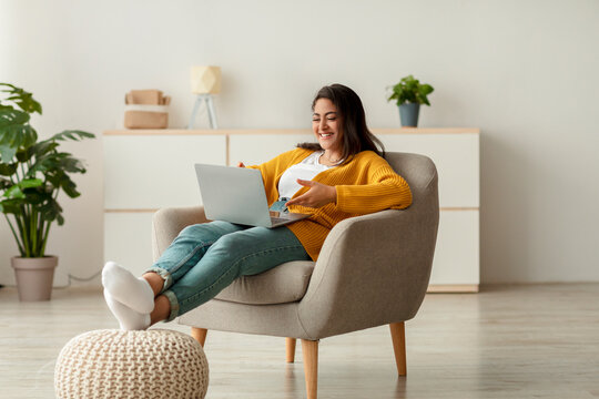 A young arab woman sits comfortably in an armchair, engaging in remote studies or video conferencing with her laptop. She enjoys the process in a bright, relaxed home environment.