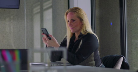 Business woman smiling while using smartphone at desk during work day in modern office setting, engaged in communication or checking messages