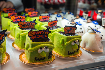Bright green square cakes styled as Frankenstein sit in a display case in Osaka, Japan. Ghost mousse cakes with blue sprinkles sit behind. Warm light and shallow depth of field.