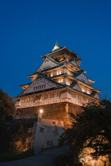 Fototapeta premium Osaka Castle in Osaka, Japan at twilight, white walls and green roofs glow, gold accents shine, soft uplighting adds drama, trees frame the scene, stone base anchors