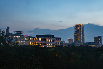 Evening cityscape in Osaka, Japan, with a lit high rise, mid rise offices, grid window lights, dark treetops in the foreground, and soft dusk clouds across the blue sky.