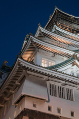 Osaka Castle in Osaka, Japan, rises at twilight, upward angled. Lit white walls, green roofs, gilded shachihoko, and lattice windows define a close night view.
