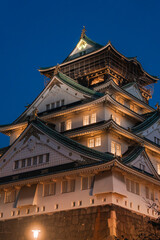 Fototapeta premium Osaka Castle in Osaka, Japan stands at dusk, green tiled roofs and gilded details lit warm, stone walls below, ornate gables and white plaster seen in soft light