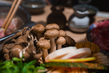 Clusters of shimeji at center with maitake, kamaboko, corn, greens, and thin meats, chopsticks and pot blur behind. Warm low light and shallow depth of field in Osaka.