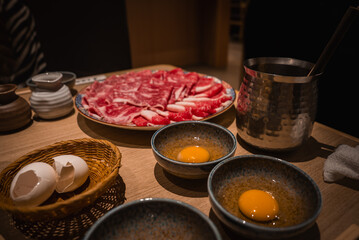 Thinly sliced marbled beef sits ready for shabu shabu or sukiyaki in Osaka, Japan. Three bowls hold raw egg yolks beside cracked shells and a metal pot ladle.
