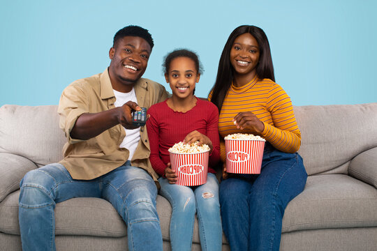 A family of three smiles while sitting on a couch with large popcorn buckets. They are ready to watch a movie together, having a great time in their lively living room.