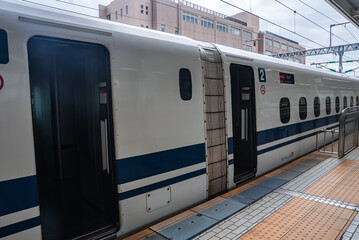 Fototapeta premium A white Shinkansen with blue stripes stops at an Osaka station platform. Doors open at car 2 under a canopy, showing aerodynamic body and tidy markings in soft daylight.