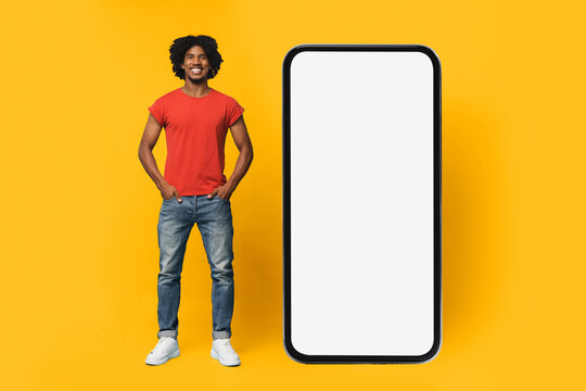 A young man with curly hair in a red shirt stands next to a tall blank smartphone on a bright yellow background. The scene emphasizes modern technology and personal style.