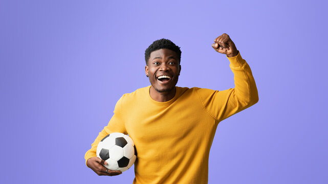 A joyful young man in a yellow sweater holds a soccer ball while raising his fist in excitement. He stands against a vibrant purple background, showcasing his enthusiasm for sports.