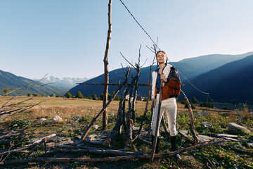 Fototapeta premium Woman portrait and traveler standing by rustic wooden fence in meadow below mountains, landscape and nature scene with clear sky, sunlight and autumn fashion jacket and boots.