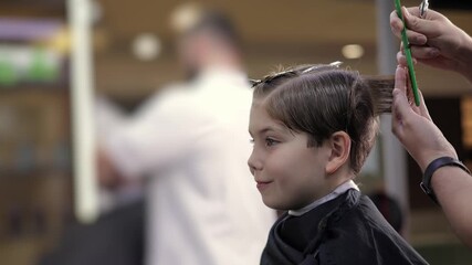 Young caucasian child receiving haircut with scissors and comb in hairstyling salon barbershop, sunlight.