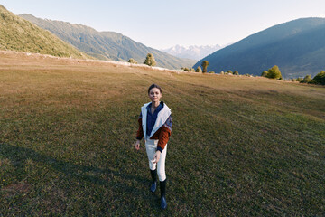 Fototapeta premium Woman walking in a meadow with mountains and expansive landscape, nature portrait of a lone hiker in open field wearing jacket and boots during clear day with golden grass and distant peaks at sunrise