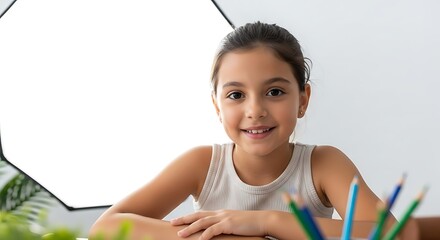 a young girl with a bright smile sits at a table holding colored pencils illuminated by soft studio lighting