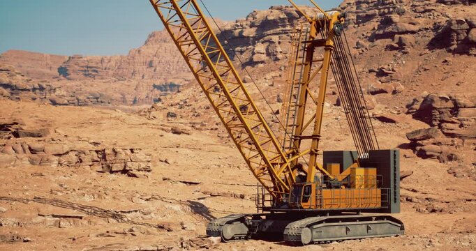 A large construction crane stands prominently in a rocky desert area, surrounded by rugged terrain. The machinery is engaged in earthmoving activities under clear blue skies during daylight.