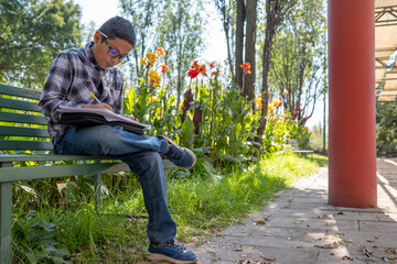 Mexican boy student wearing glasses writing homework outdoors in park