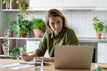 Stressed middle aged woman writing down budget plan and utility bills while sitting in kitchen using laptop. Independent female homeowner plans monthly expenses, checking financial stats on computer.