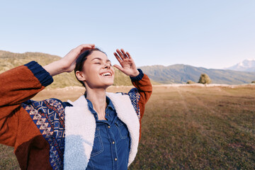 Fototapeta premium Woman smiling with eyes closed outdoors in countryside, hands raised enjoying nature and warm sunshine, happiness and freedom in casual jacket and denim, relaxed joyful portrait in meadow.