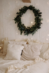 Christmas wreath above bed in a beige bedroom with textured white wall. Warm lights, ruffled pillows and soft bedding create a cozy holiday interior for seasonal decor and home comfort.