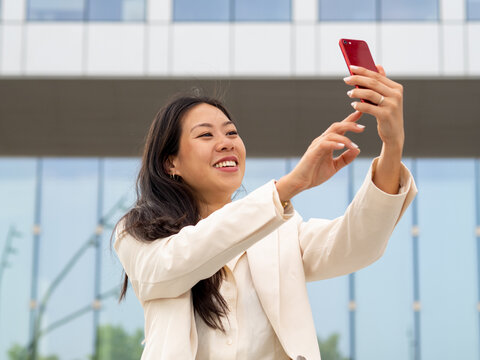 Young Asian businesswoman taking a selfie with her smartphone, smiling confidently while standing outdoors in a modern urban setting.