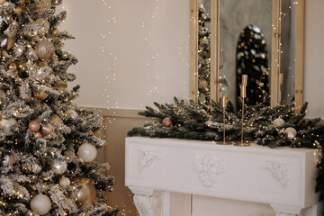 Frosted Christmas tree with gold ornaments stands beside a white classic mantel decorated with evergreen garland, candles and warm lights in a soft interior. Suitable for holiday themes, decor. 