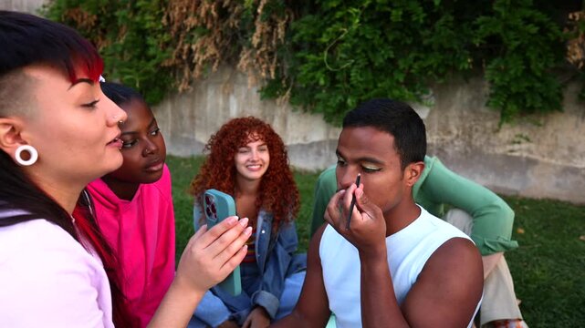 Diverse group of young friends laughing and having fun together outdoors in a park while a man applies eyeliner and eyeshadow, embracing self expression and community without judgment