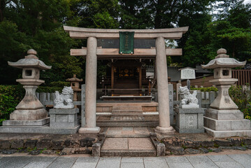 Stone torii stands with komainu and lanterns at a Kyoto shrine. Short steps lead to a wooden honden. Overcast light softens weathered stone and neat paving.