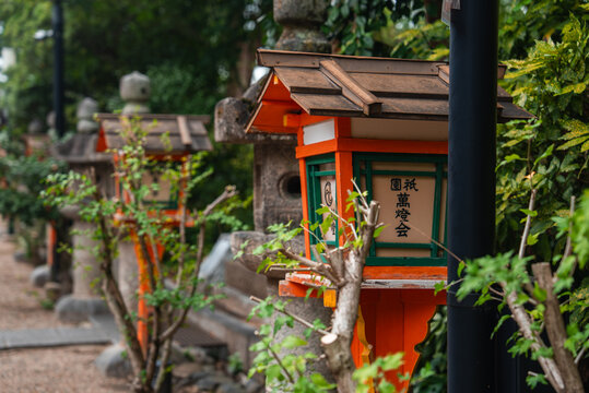 Traditional stone lanterns and orange and green lamps line a gravel path in Kyoto, with kanji signage visible. Soft overcast light and shallow focus highlight wood and stone.