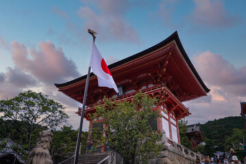 A vermilion gatehouse at Kiyomizu dera in Kyoto, Japan, stands with a flag, lion dog statues, and...