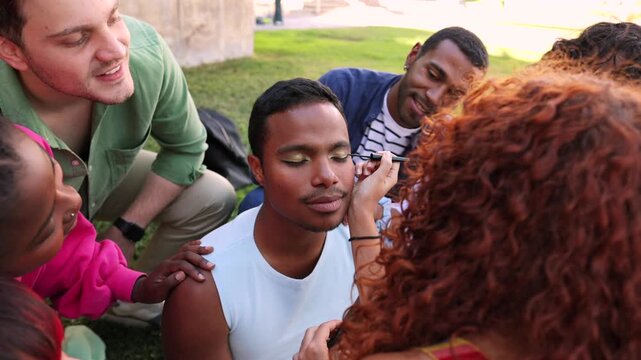 Young multicultural friends having fun while a woman applies makeup to a man's eye, celebrating friendship, diversity, and self expression in a park with smiles and support from the group
