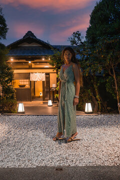 A woman pauses in a gravel forecourt at a machiya house in Kyoto at dusk. Lanterns light a tiled roof, wooden beams, noren, and trees as a pink and purple sunset colors the scene.