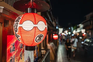 Fotobehang Smal steegje A red and white chochin with bold kanji hangs by a wooden shop in Kyoto, with machiya facades and lanterns along a narrow lane as people stroll in soft focus.  © Aerial Film Studio