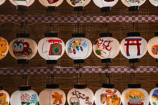 Rows of hand painted lanterns show torii, koi, masks, and calligraphy. Checkered trim and bamboo backdrop suggest a matsuri in Kyoto, Japan. Warm even light shows texture.