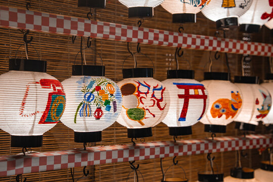 Rows of Japanese paper lanterns hang in Kyoto, Japan, with torii, kabuki mask, koi, and motifs. Warm even lighting shows washi texture and brushwork in close view.