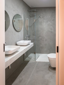 A contemporary bathroom features microcement walls and floors, double sinks, and a glass shower, embracing a modern minimalist aesthetic that highlights durability and seamless design.