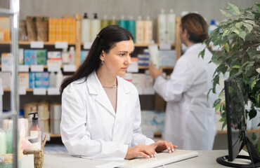 Female pharmacist is concentrating on working at the computer, she enters information about new pills, against the background of a male pharmacist checking the number of packages on the shelves