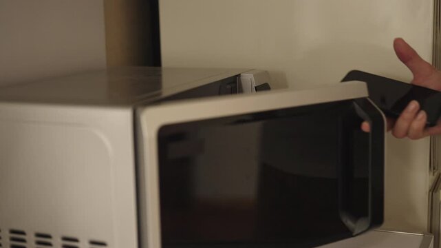 Close-up of unrecognizable person placing smartphone inside microwave oven, illustrating danger of misuse, technological ignorance, risk of damaging electronic devices. Shooting in slow motion.