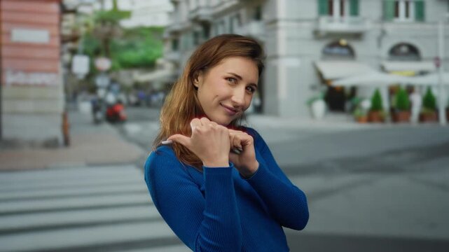 Woman gestures with thumbs backwards on a city street, wearing a blue sweater, depicting confidence and playfulness outdoors during the day.