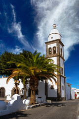 A stunning view of the historic church in Arrecife, Lanzarote, surrounded by tall palm trees. The bright blue sky enhances the picturesque scene, capturing the essence of this charming location