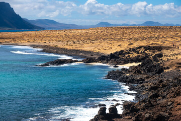 Rugged volcanic coastline of La Graciosa features turquoise waters meeting black rocks. Majestic cliffs rise in the distance under blue skies. Visitors walk along the sandy path