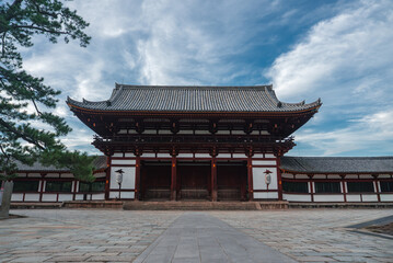 A grand wooden gate with red pillars and white walls stands in Nara, Japan. A tiled roof and eaves frame a stone forecourt leading to the entrance in daylight. © Aerial Film Studio