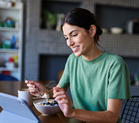 Portrait of a young woman using tablet or laptop computer, having online meeting or browsing internet, drinking coffee eating breakfast, drinking juice  in the kitchen at home