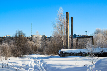Grim concrete skeletons of an unfinished building rise amidst a serene snowy winter landscape under a bright blue sky. A frosty path leads onward