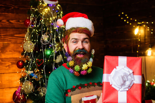 Winter holidays celebration. Christmas and New Year sales. Smiling man with decorated beard holds Christmas gift box. Bearded man with dyed hair in Santa hat holds Christmas present. Delivery service.