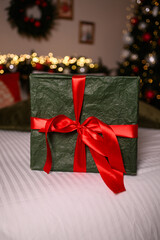 A green wrapped gift box with a bright red ribbon sits on a white bedspread, surrounded by soft Christmas lights and festive holiday decor in the blurred background.