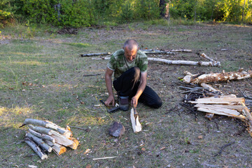 A man is chopping firewood in the forest with an axe. He is a middle-aged man with short hair. A dog is sitting on the grass nearby.