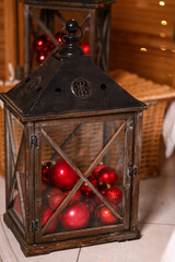 Vintage wooden lantern filled with red Christmas ornaments standing indoors as part of festive holiday d cor. Warm lights and wicker elements in the background create a cozy seasonal atmosphere.